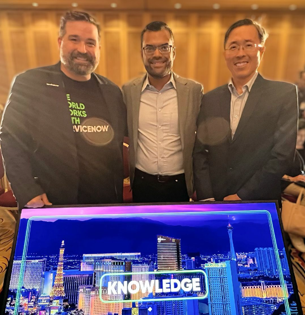 Three smiling men stand together at a ServiceNow Knowledge conference, with a screen showing a Las Vegas skyline behind them.